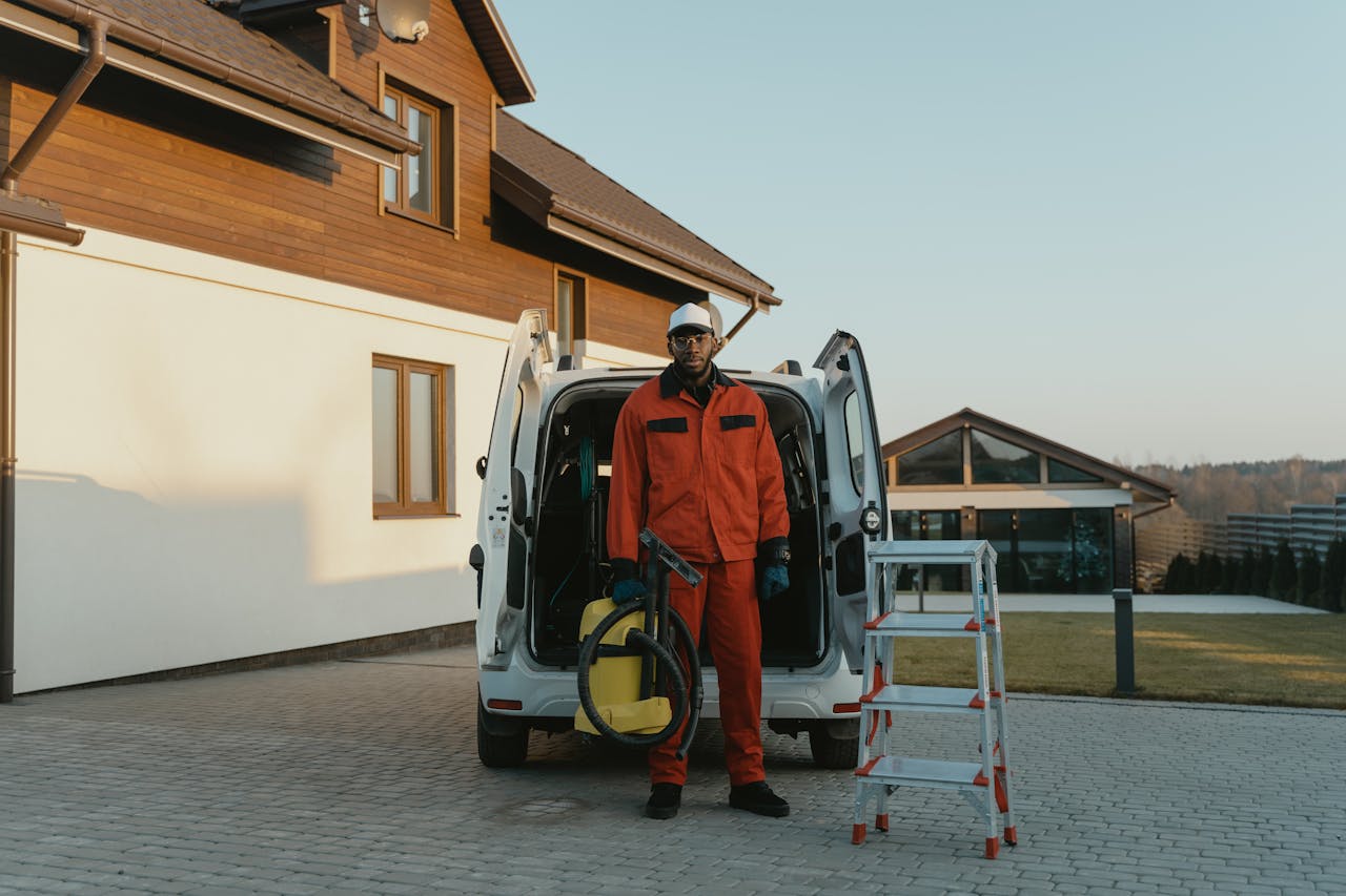 A male worker in red coveralls with a vacuum cleaner beside a van on a sunny day.