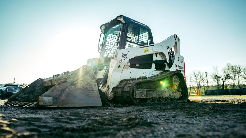 A skid steer loader on a construction site with bright sunlight in the background.